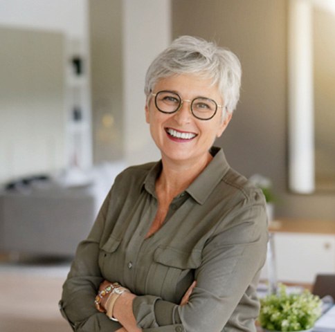 Senior woman with glasses standing in room smiling with arms folded